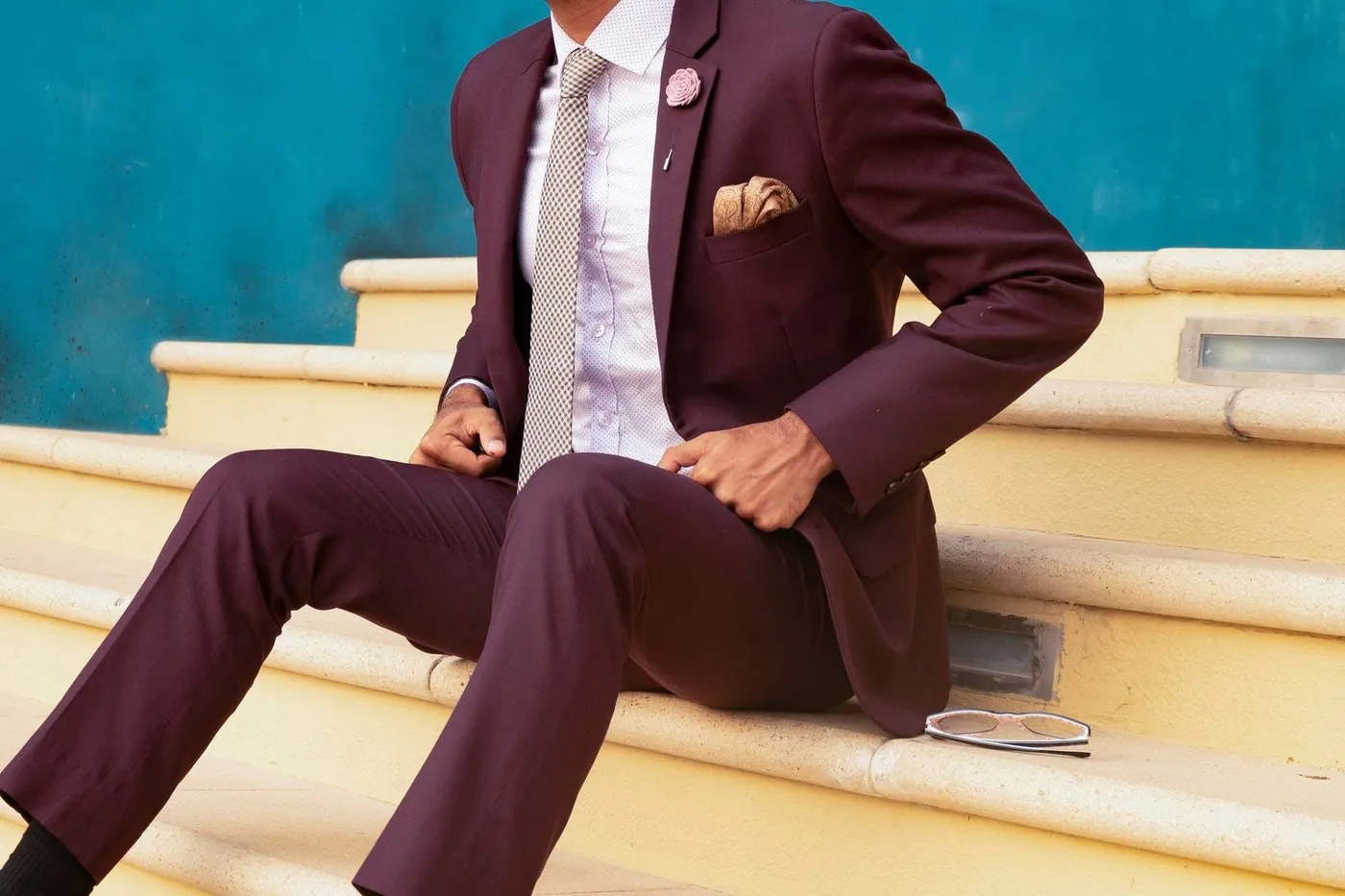 A person wearing a burgundy suit, dress shirt, tie, and pocket square while seated on steps