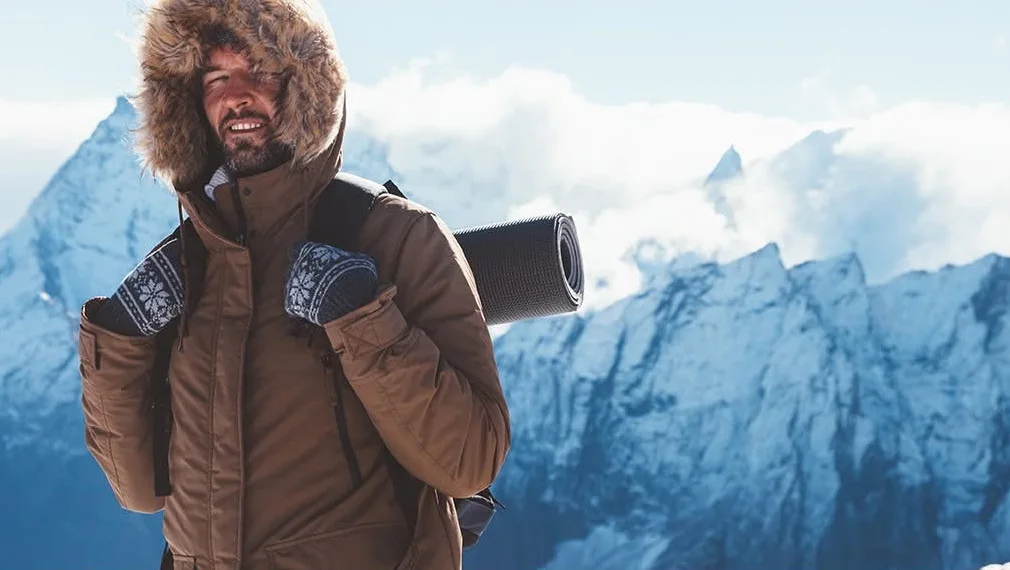 A man in a brown hooded parka carrying a backpack in front of snowy mountains